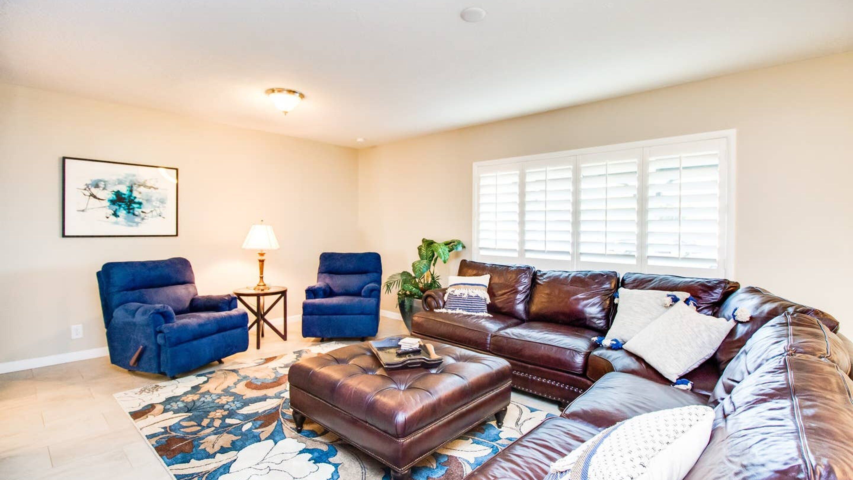 A living room with a brown leather sofa, two blue armchairs, a small side table with a lamp, a large window with blinds, and a framed abstract painting on the wall.