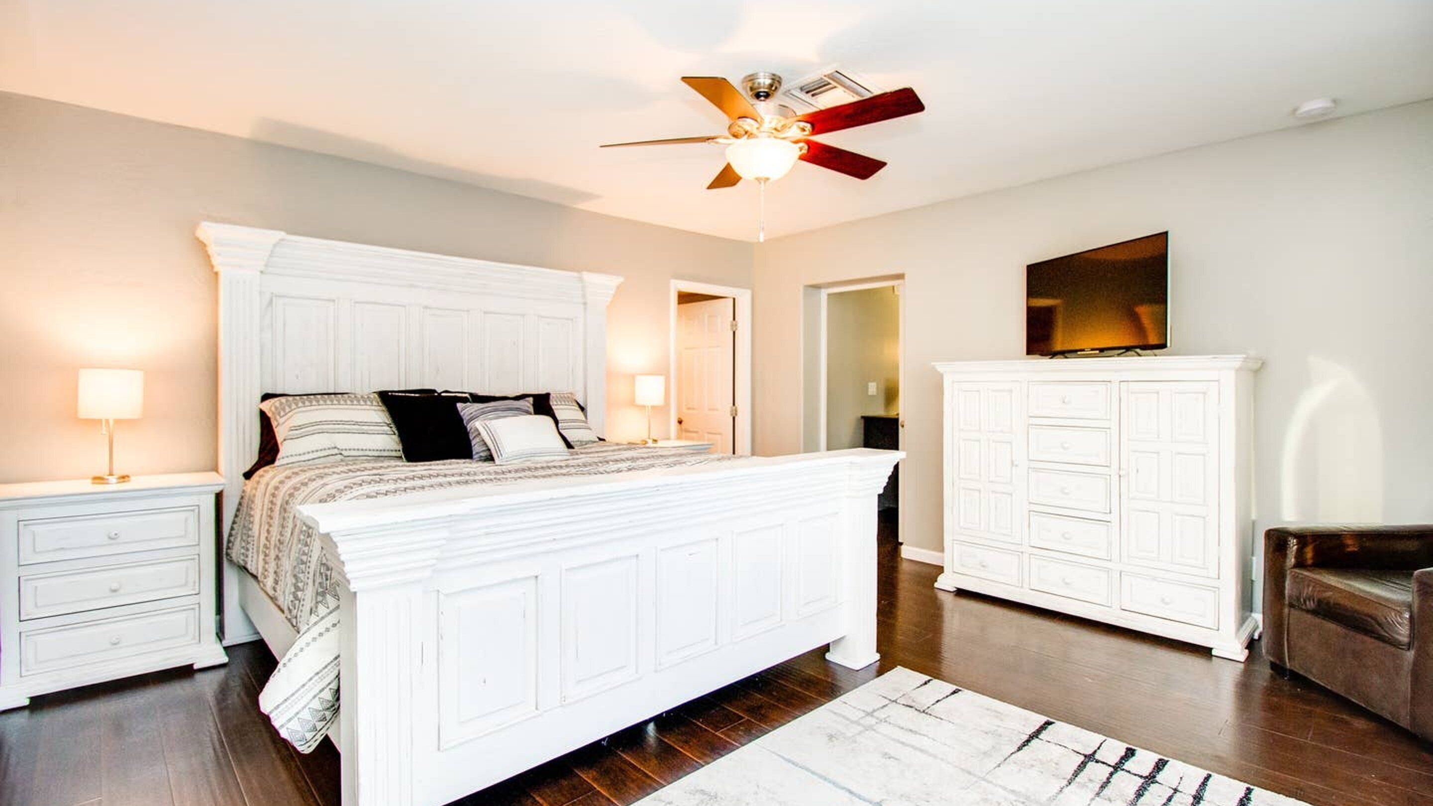 A bedroom with a large bed, white nightstands, a ceiling fan, a wardrobe, and a chair.
