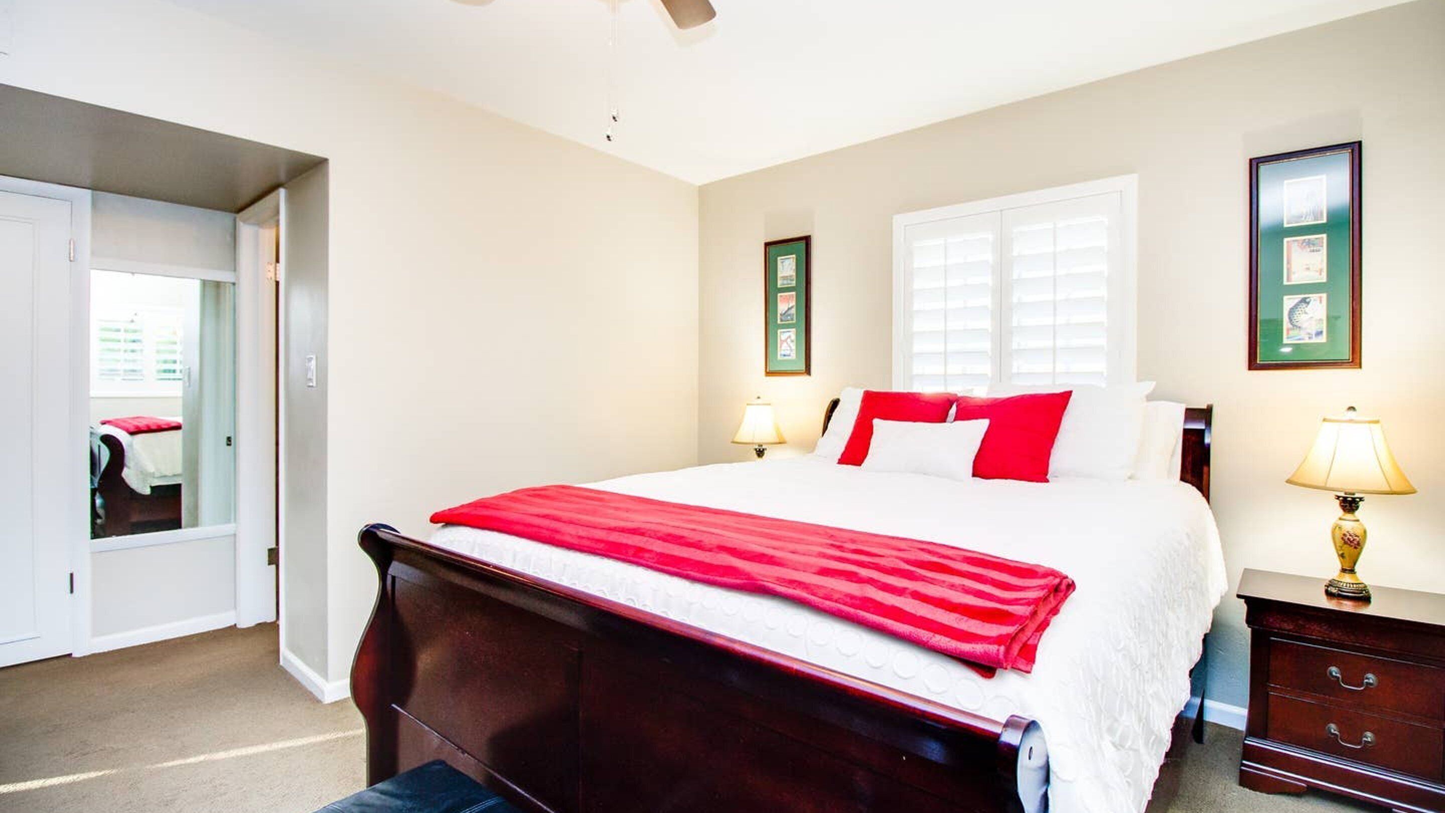 A bedroom with a large bed, two bedside tables with lamps, a window with blinds, and a door leading to another room.