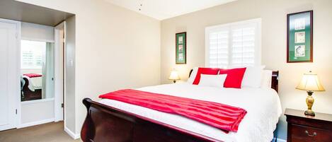 A bedroom with a large bed, two bedside tables with lamps, a window with blinds, and a door leading to another room.