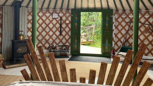 A yurt with a traditional design, featuring a central wooden structure, a cozy seating area with cushions, and a view of the outdoors.