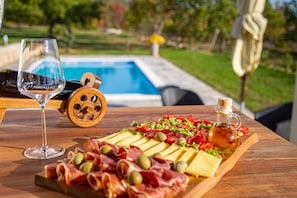 Une table en bois sur laquelle on aperçoit une planche à fromages, des olives et de la charcuterie. On y voit également un verre à vin et une bouteille d’huile d’olive.
