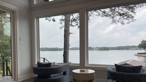 Une pièce dotée de grandes fenêtres donnant sur un lac, avec deux fauteuils bleus et une table d’appoint en bois.