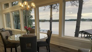 Un coin repas avec une table ronde, des chaises et un lustre. De grandes fenêtres offrent une vue sur un lac et des arbres.