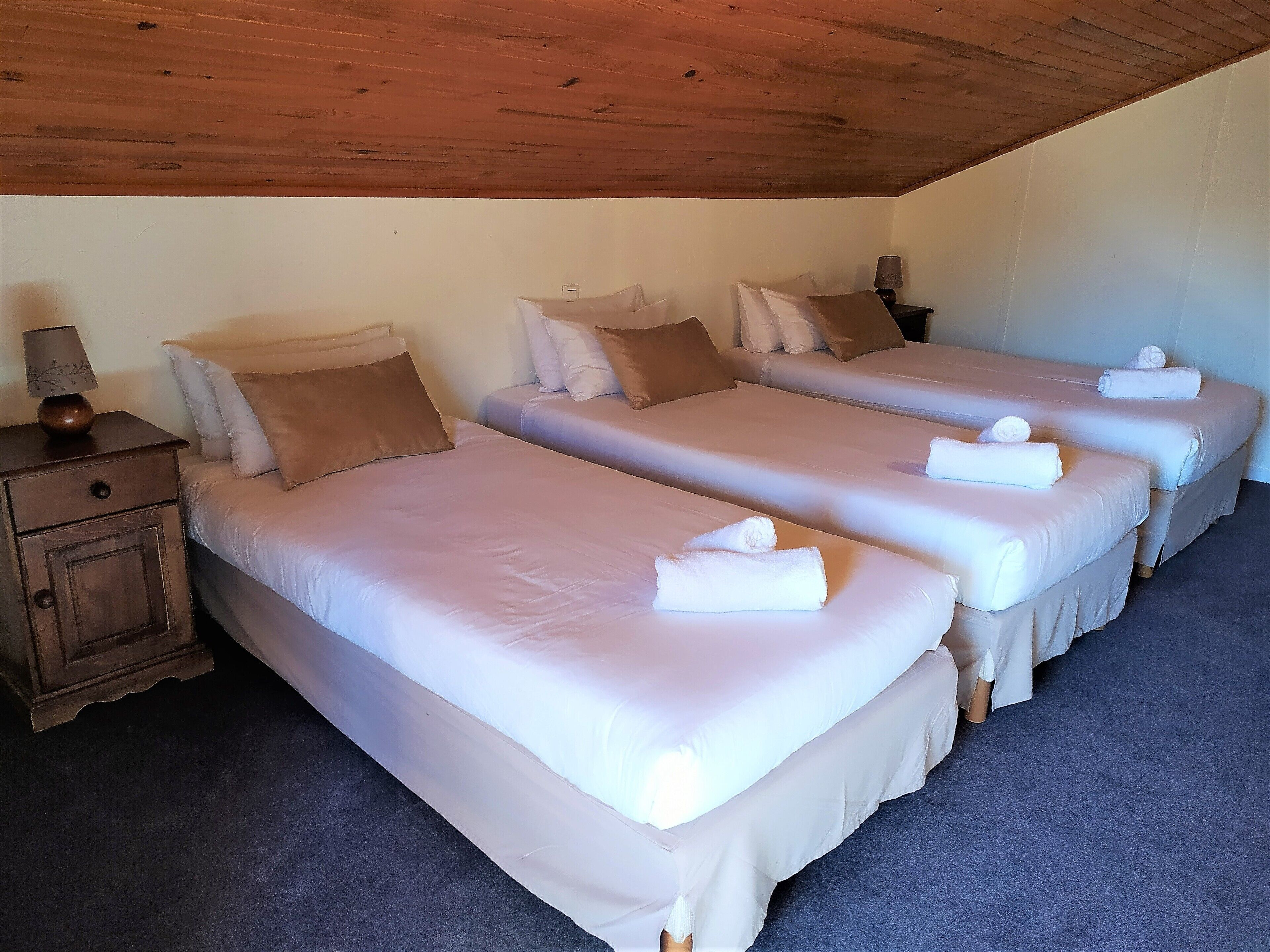 Habitación de hotel con tres camas, cada una con sábanas blancas y almohadas marrones. Hay una mesita de noche de madera con una lámpara y un pequeño armario.