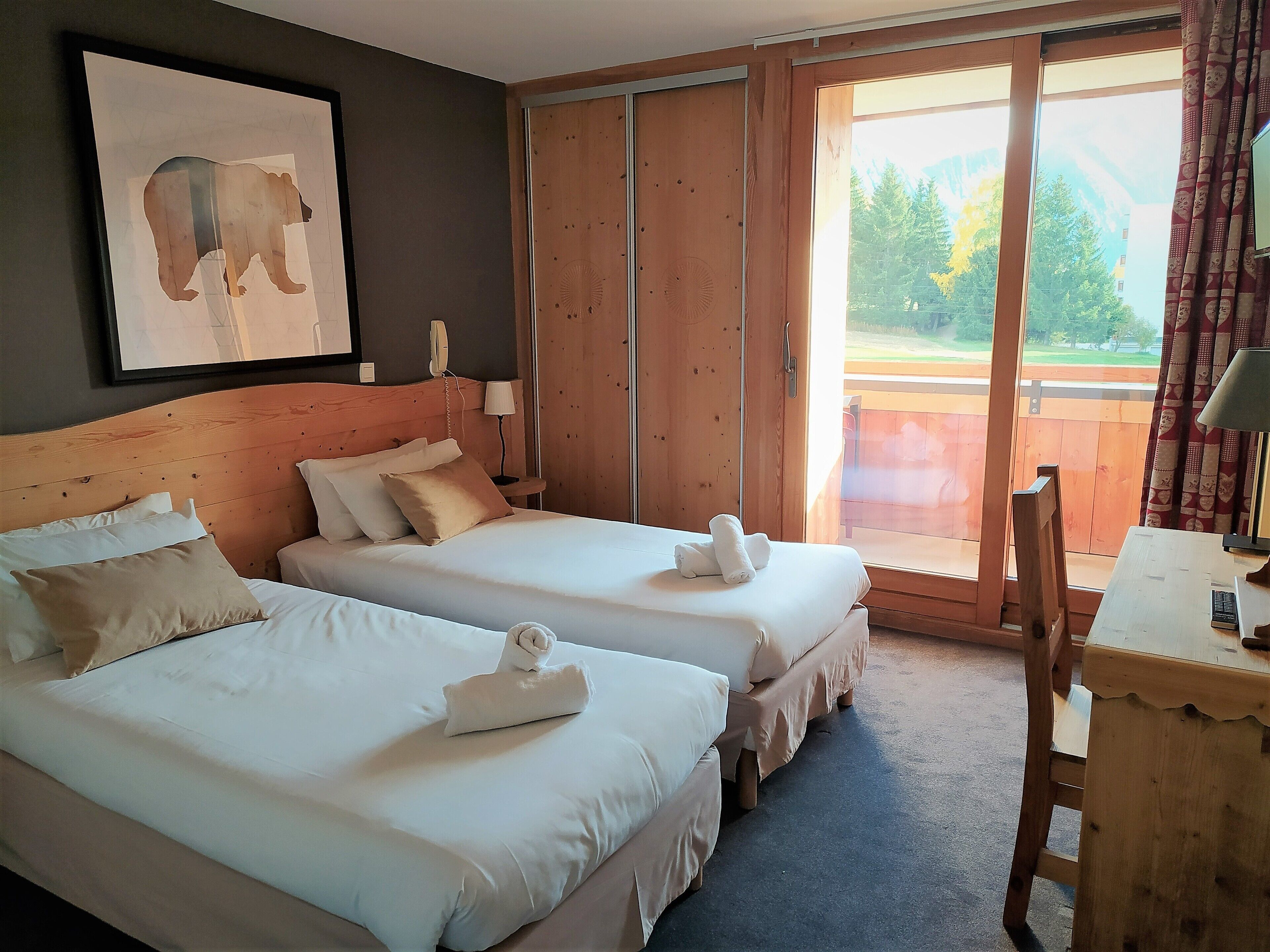 A hotel room with two beds, a wooden headboard, a painting of an elephant, a telephone, and a view of a balcony with a wooden railing.