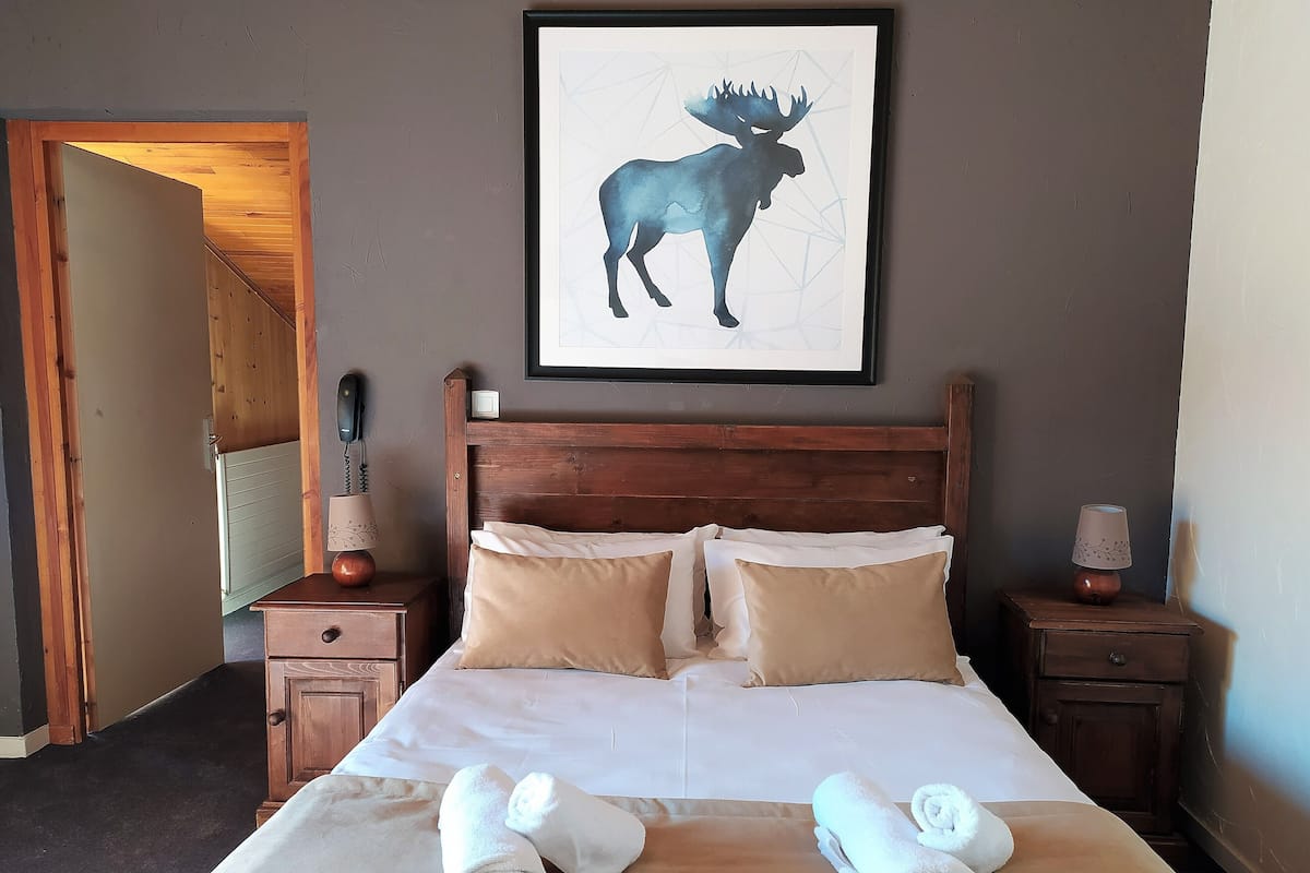 A hotel room with a wooden headboard, a framed moose artwork, a bed with white linens, two bedside tables with lamps, and rolled towels on the bed.