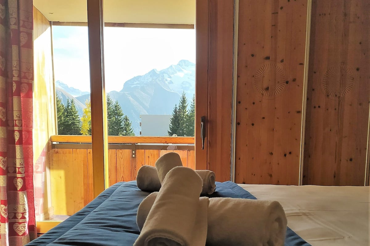 A bedroom with a bed, a view of mountains, and a balcony with wooden railings.