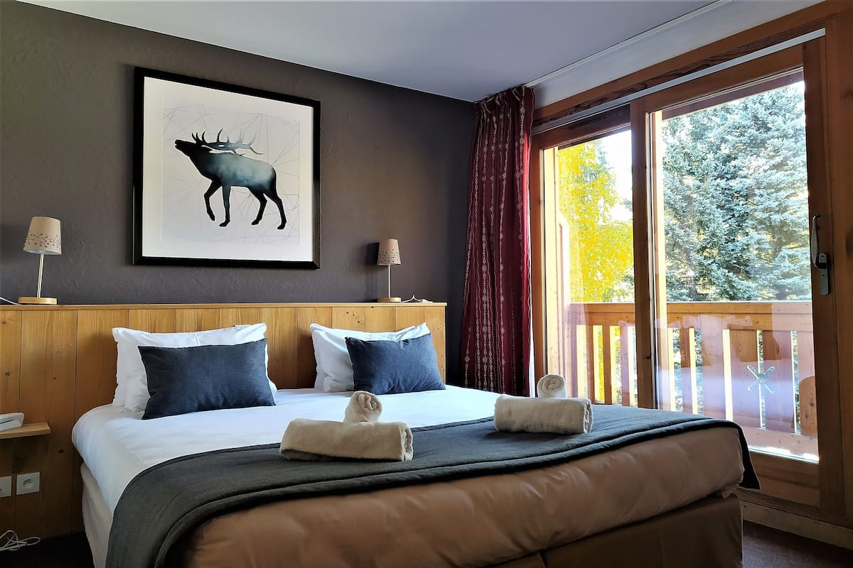 A hotel room with a large bed, two pillows, a framed artwork of a moose, a wooden headboard, a nightstand, a lamp, and a view of a deck with a railing.