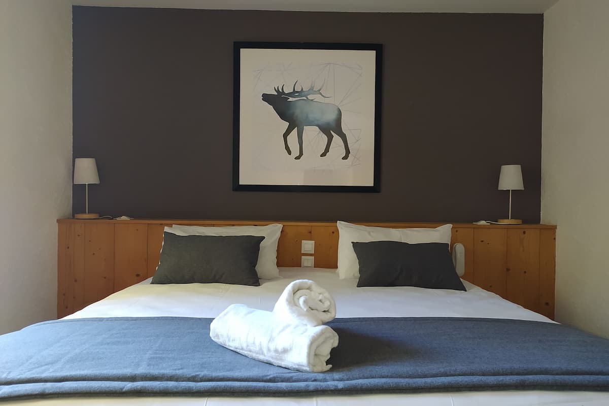 A neatly made bed with a blue blanket and a rolled towel, two pillows, a wooden headboard, and a framed moose artwork on the wall.