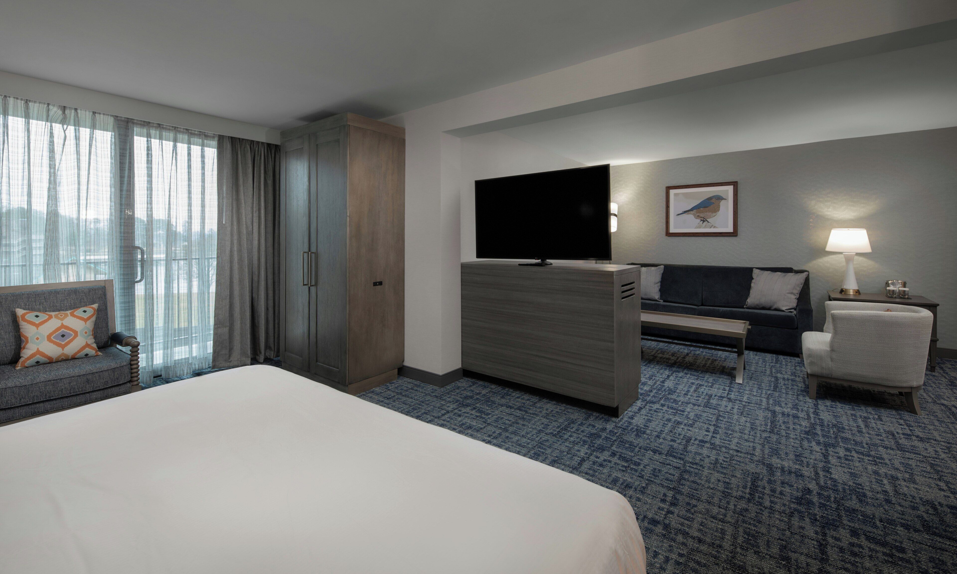 A hotel room with a bed, a TV, a sofa, a chair, a desk, and a lamp.