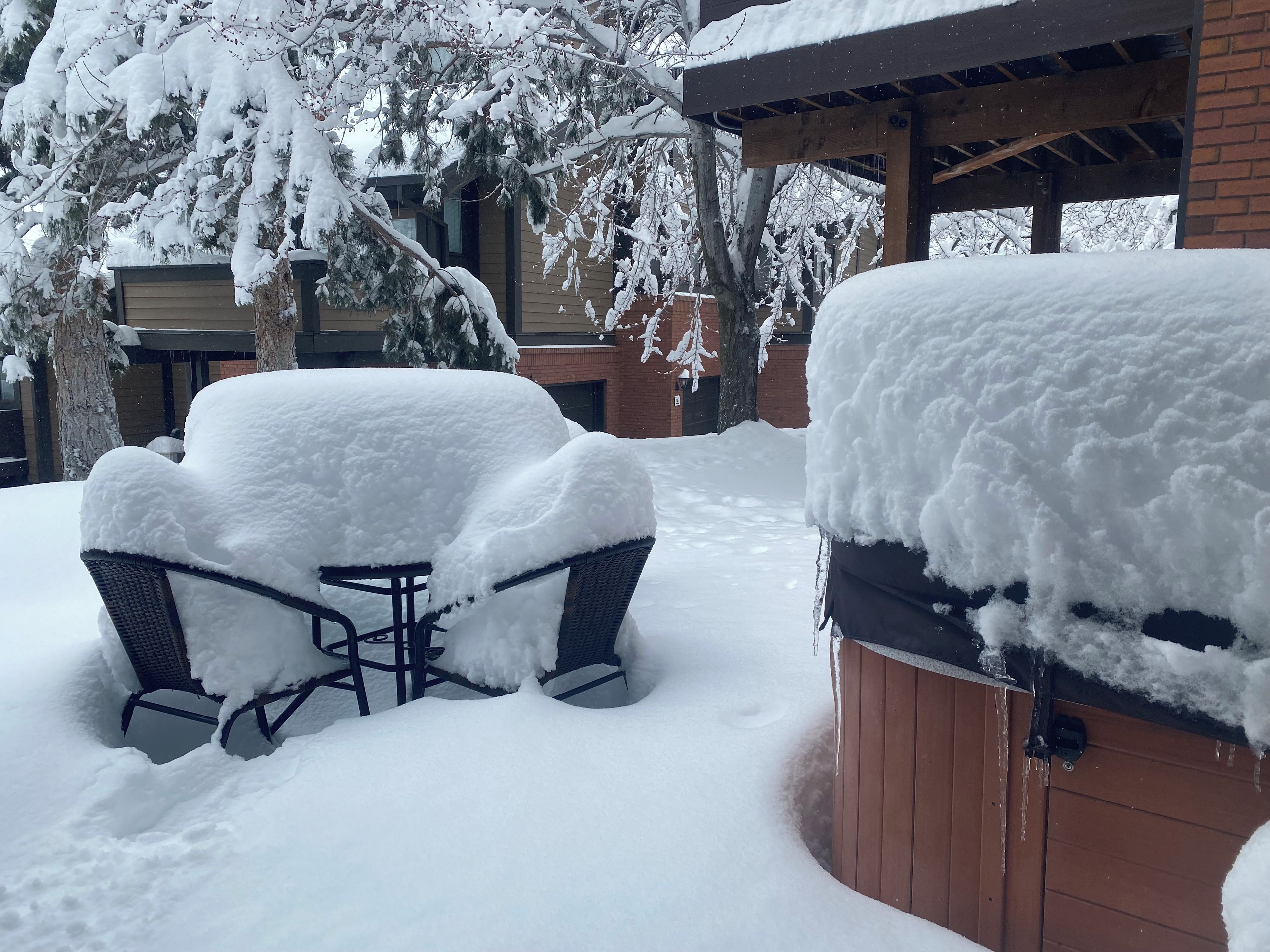 Ein mit Schnee bedeckter Außenbereich mit Sitzgelegenheiten, einem Tisch, Stühlen, einer Holzterrasse und einem Gebäude im Hintergrund.