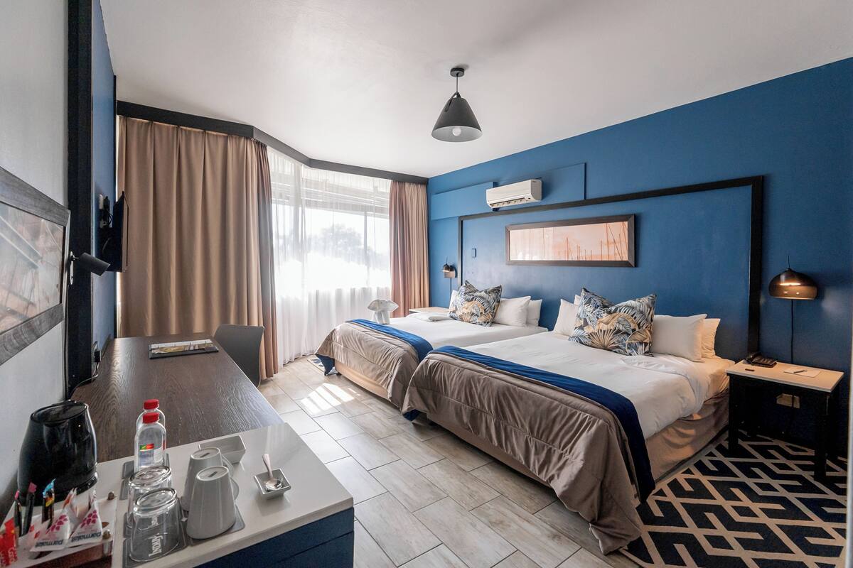 A hotel room with two beds, a desk with a coffee maker, a lamp, and a window with curtains.
