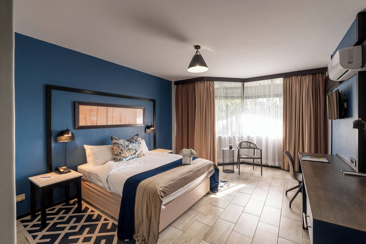 A hotel room with a bed, desk, chair, and a large window with curtains.