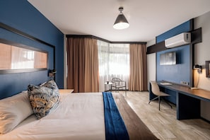 A hotel room with a bed, a desk, a chair, a TV, and an air conditioning unit.