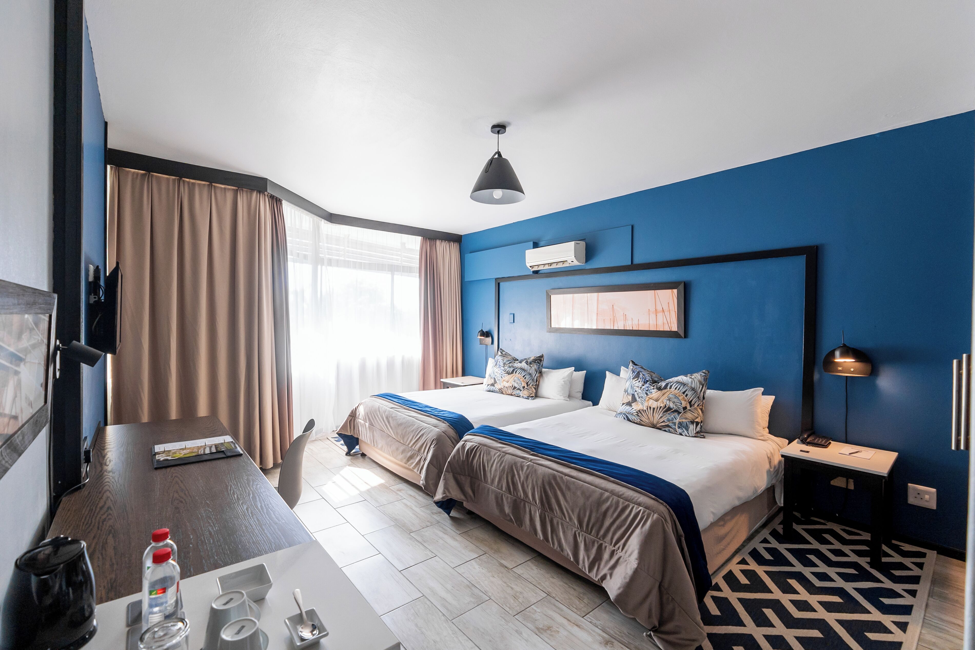 Habitación de hotel con dos camas, un escritorio, una silla, una lámpara y un ventanal con cortinas.