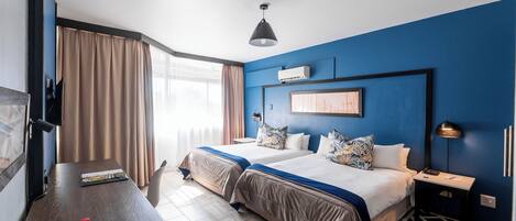 Habitación de hotel con dos camas, un escritorio, una silla, una lámpara y un ventanal con cortinas.