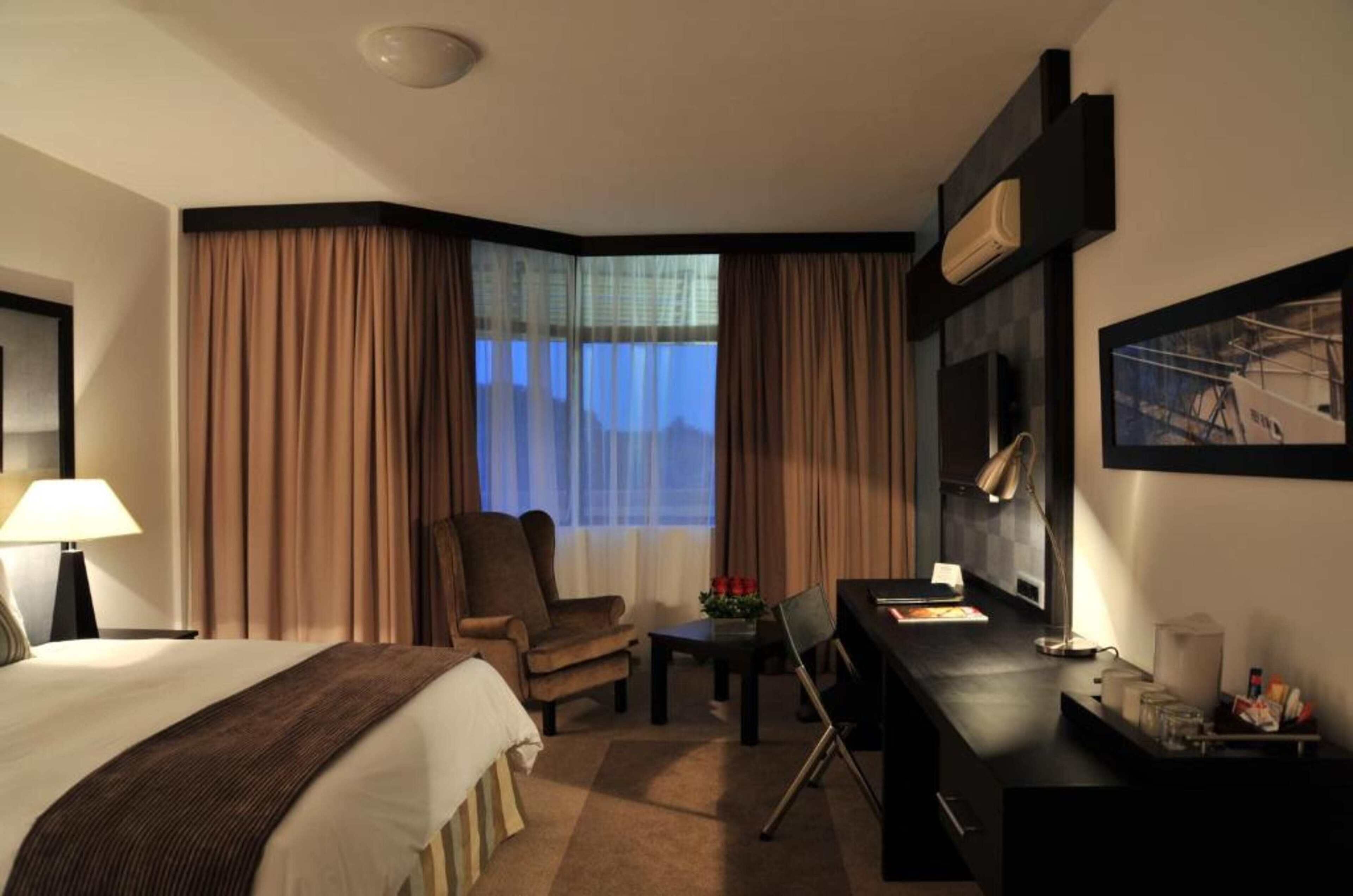 Una habitación de hotel con cama, escritorio, silla, lámpara, ventana con cortinas e imagen enmarcada en la pared.