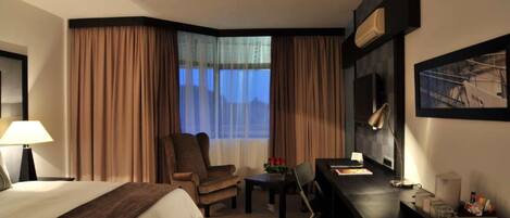 Una habitación de hotel con cama, escritorio, silla, lámpara, ventana con cortinas e imagen enmarcada en la pared.