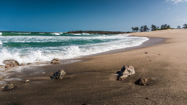 Une plage avec des vagues, une rive sablonneuse et une jetée qui s’étend au loin.