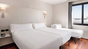 A hotel room with two beds, a bedside table, a lamp, and a window with a view of buildings.