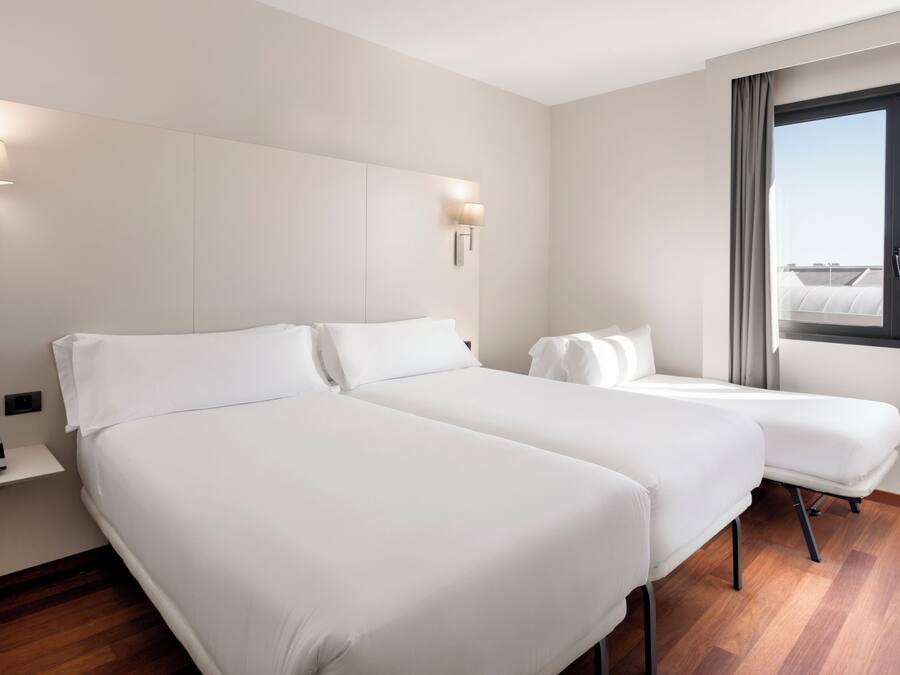 Habitación de hotel con dos camas, una mesita de noche, una lámpara y una ventana con vistas a edificios.