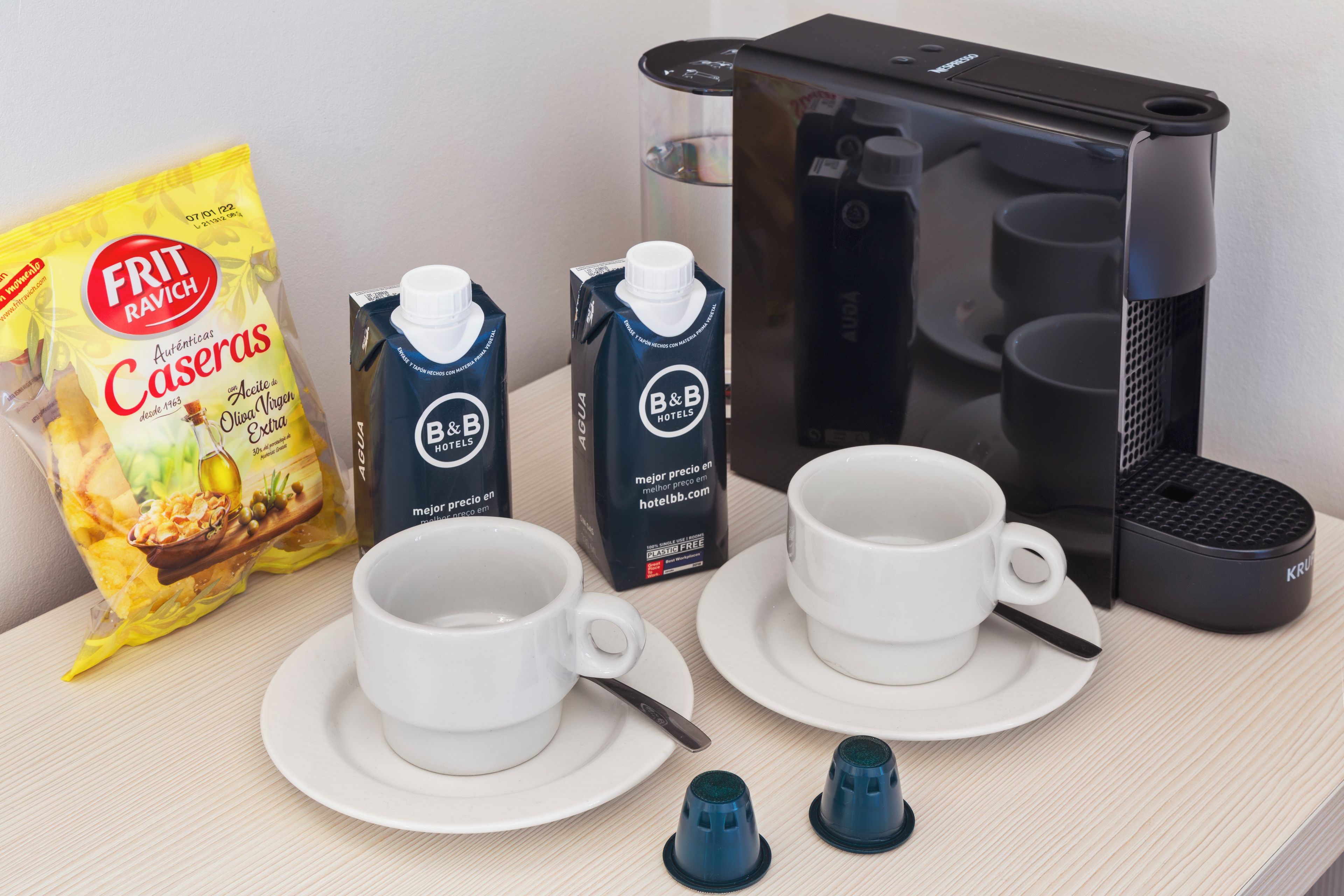 Ein Frühstücksarrangement mit zwei weißen Tassen, einer Tüte Frit Ravich Caseras Chips, zwei Milchkartons von B&B Hotels und einer Kaffeemaschine.