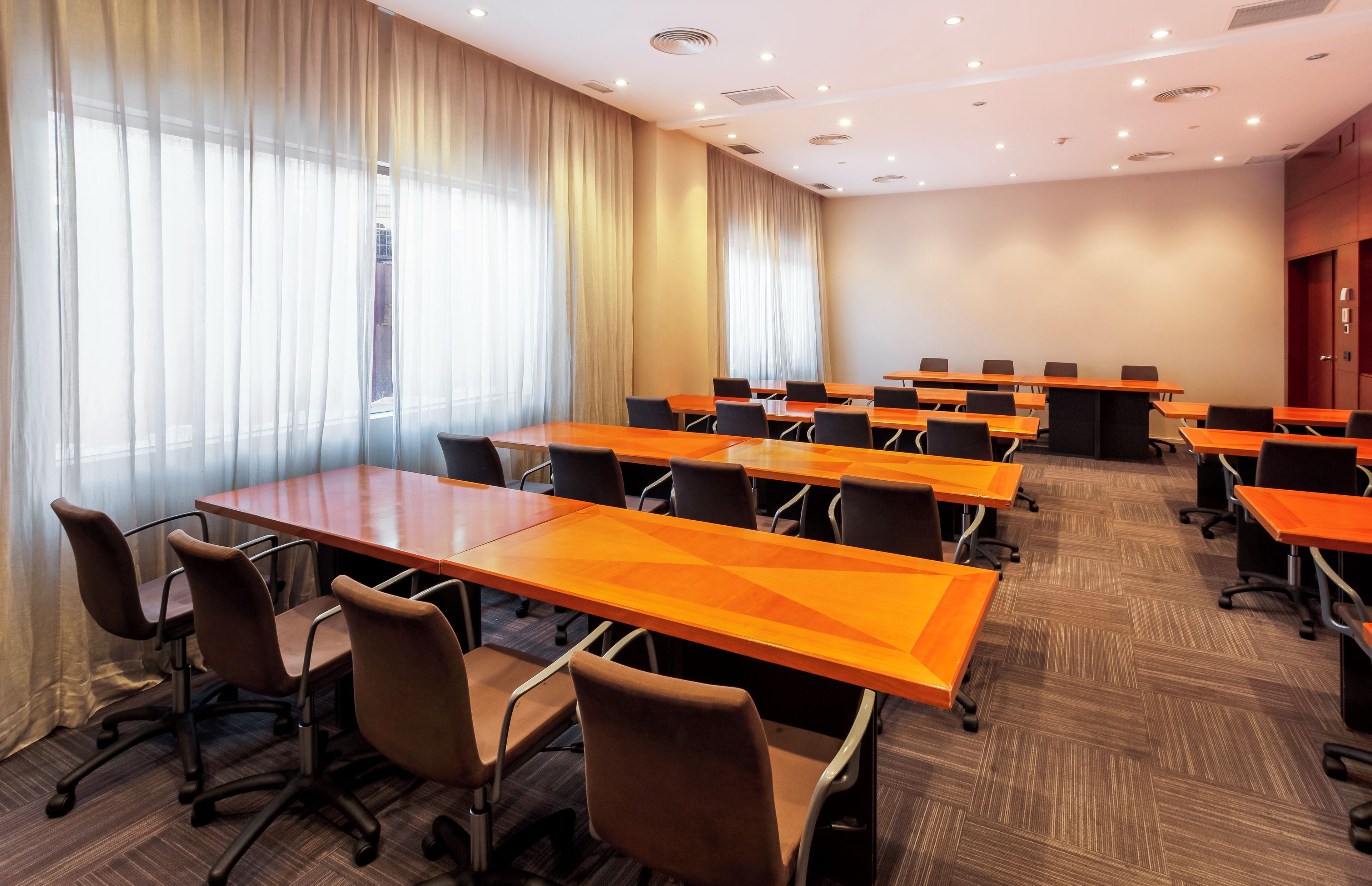 Un salón de conferencias con varias mesas y sillas, ventanas amplias con cortinas y piso alfombrado.
