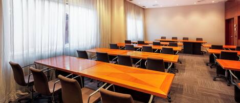 Une salle de conférence dotée de plusieurs tables et chaises, de grandes fenêtres avec des rideaux et d’un sol recouvert de moquette.