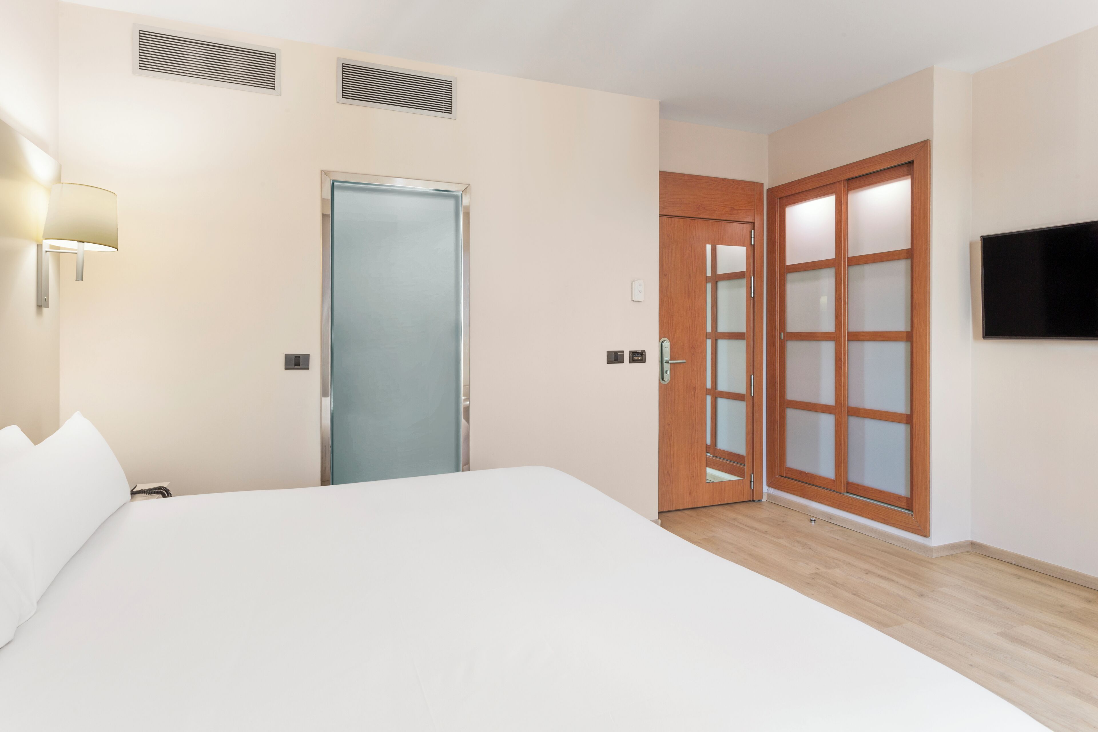 Habitació d'hotel amb llit, una porta amb panells de vidre mat, televisió penjada a la paret i làmpada fixada a la paret.