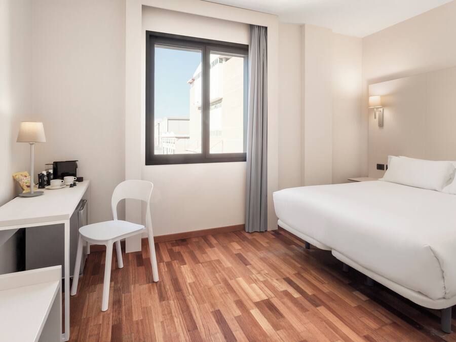 A hotel room with a bed, a desk with a chair, a lamp, and a window with curtains.