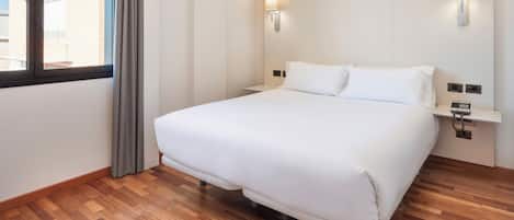 A hotel room with a large bed, wooden flooring, two wall-mounted light fixtures, and a window with curtains.