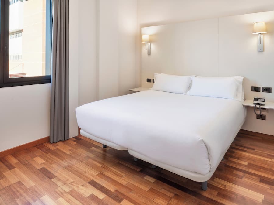 Una habitación de hotel con una cama grande, suelo de madera, dos lámparas fijadas a la pared y un ventanal con cortinas.