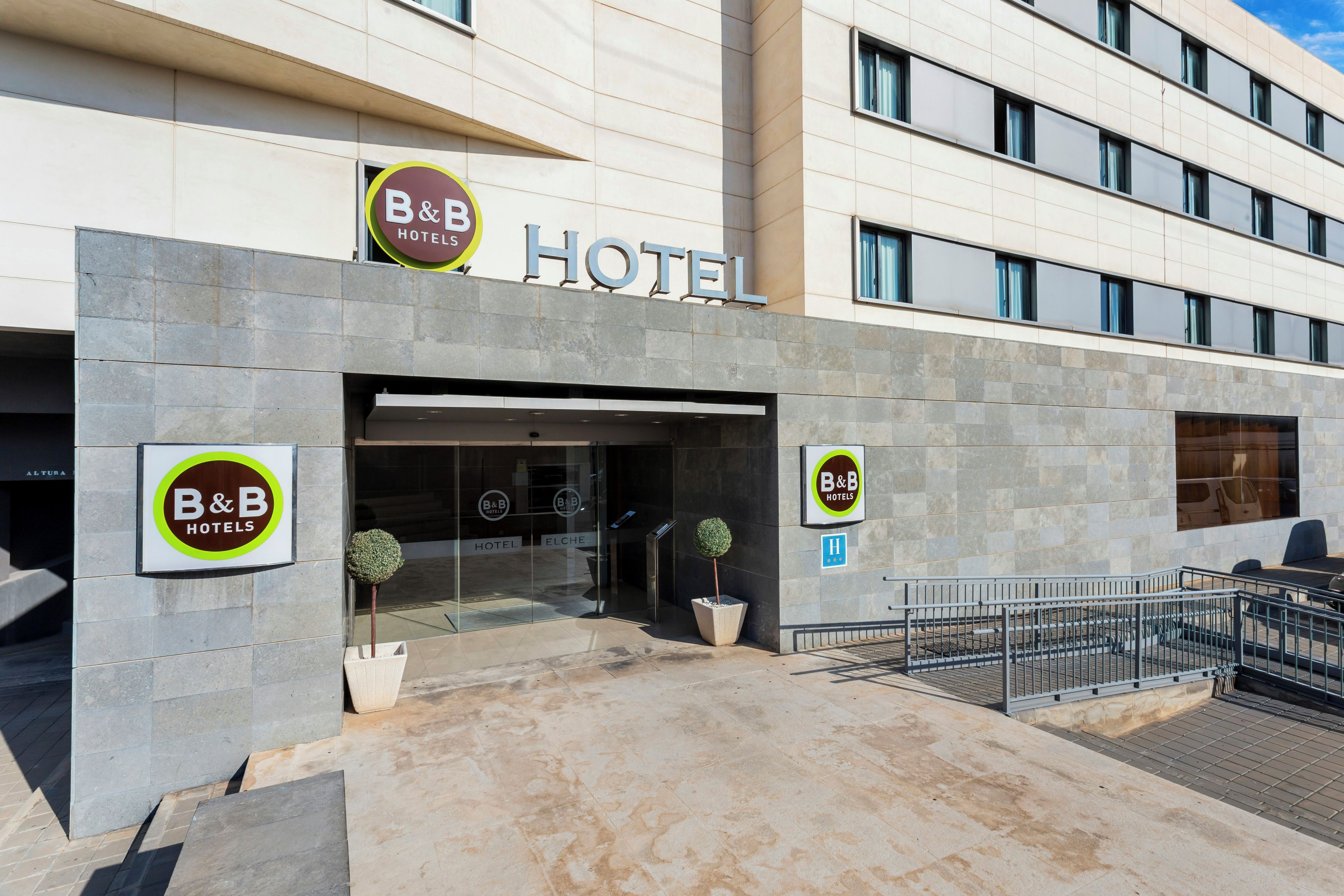 Entrada de un hotel B&B con fachada moderna, puertas de vidrio y una pequeña planta en maceta.