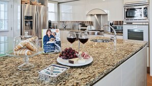 A modern kitchen with stainless steel appliances, a granite countertop, and a dining area with a glass display case, a wine glass, and a cheese board.