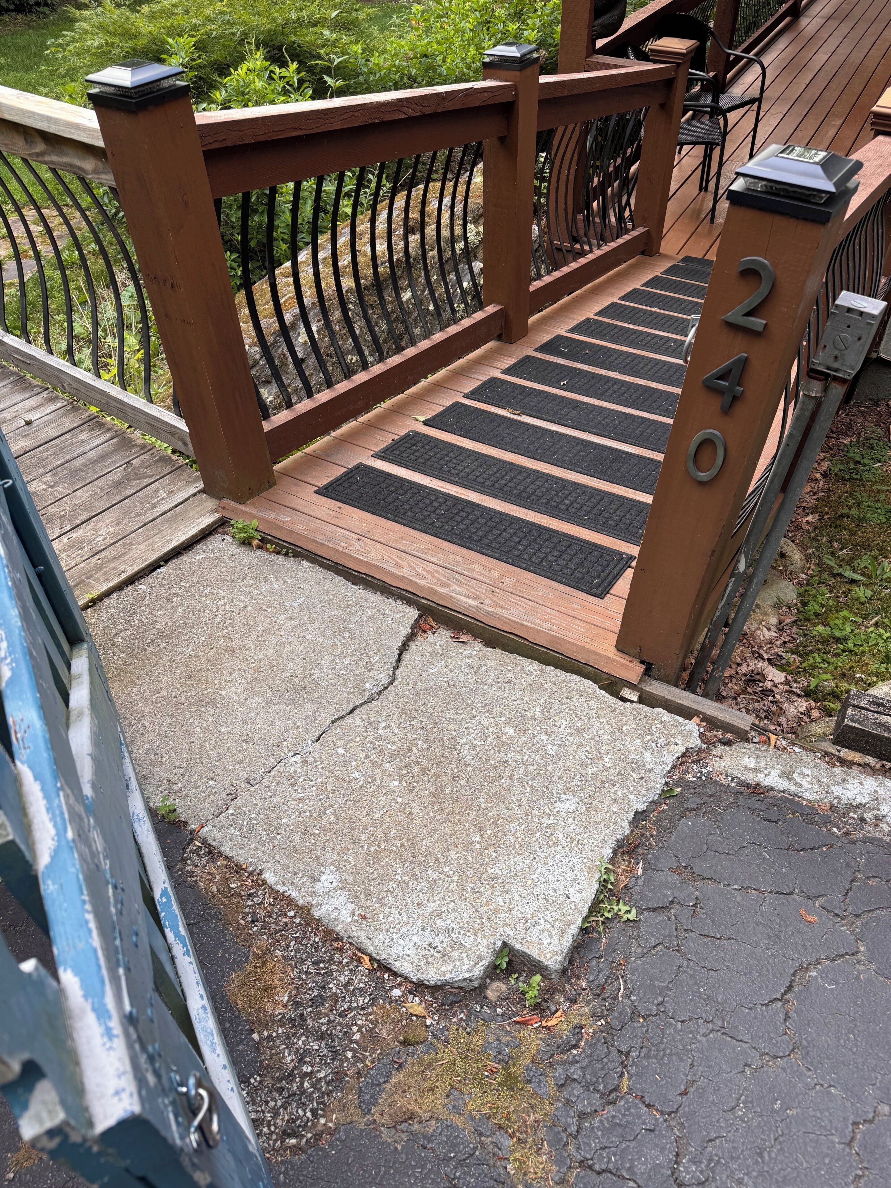 A wooden deck with a metal railing, a concrete path with black rubber mats, and a metal gate with the number 2400.