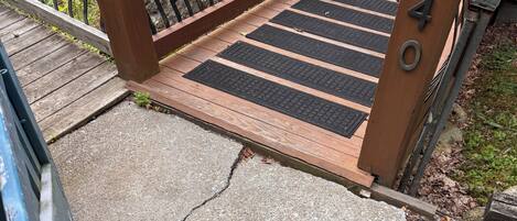 A wooden deck with a metal railing, a concrete path with black rubber mats, and a metal gate with the number 2400.