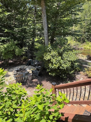 A wooden deck with a stone fire pit and chairs, surrounded by dense greenery and trees.