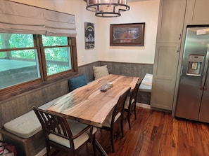 A dining area with a wooden table, chairs, a bench, a refrigerator, and a window with blinds.