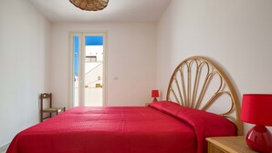 Un dormitorio con una cama, una mesita de noche de madera con una lámpara roja, una silla de mimbre y vista a un edificio por una puerta.