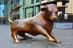 Une statue de taureau en bronze, située dans un environnement urbain, avec des bâtiments modernes en arrière-plan.