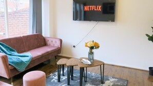 Ein Wohnzimmer mit einem rosafarbenen Sofa, einem Couchtisch mit Blumenvase und einem an der Wand befestigten Fernseher, der das Netflix-Logo zeigt.