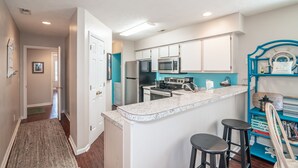 A kitchen with white cabinets, a stainless steel refrigerator, a microwave, and a stove. There are two black barstools and a countertop. - TB 12A First Floor 3BR/3BA with great view of golf course w/no stairs (PAWLEYS ISLAND)