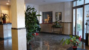 Eine Lobby mit Marmorboden, einer großen Topfpflanze, einem kleinen Tisch mit einer Vase und einem Fenster mit Blick auf ein Gebäude.