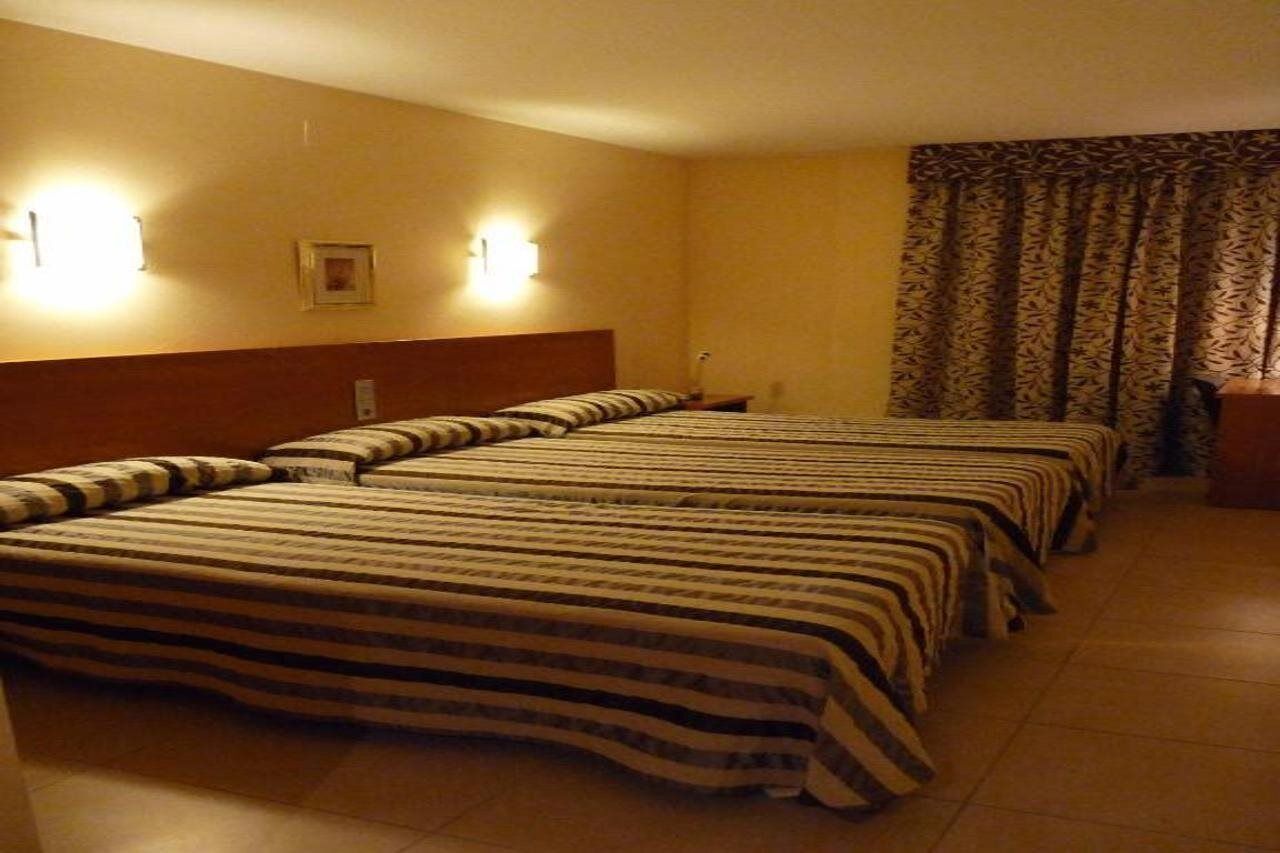 Ein Hotelzimmer mit drei Betten, einem Holzkopfteil, zwei Wandleuchten, einem gemusterten Vorhang und einem gerahmten Bild an der Wand.