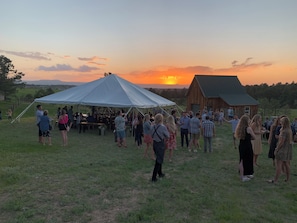 A group of people gathered under a large tent in a grassy field with a barn in the background during sunset.
