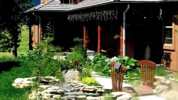 Ein zweistöckiges Haus mit Veranda, umgeben von einem Garten mit einem Steinbrunnen und Stühlen.