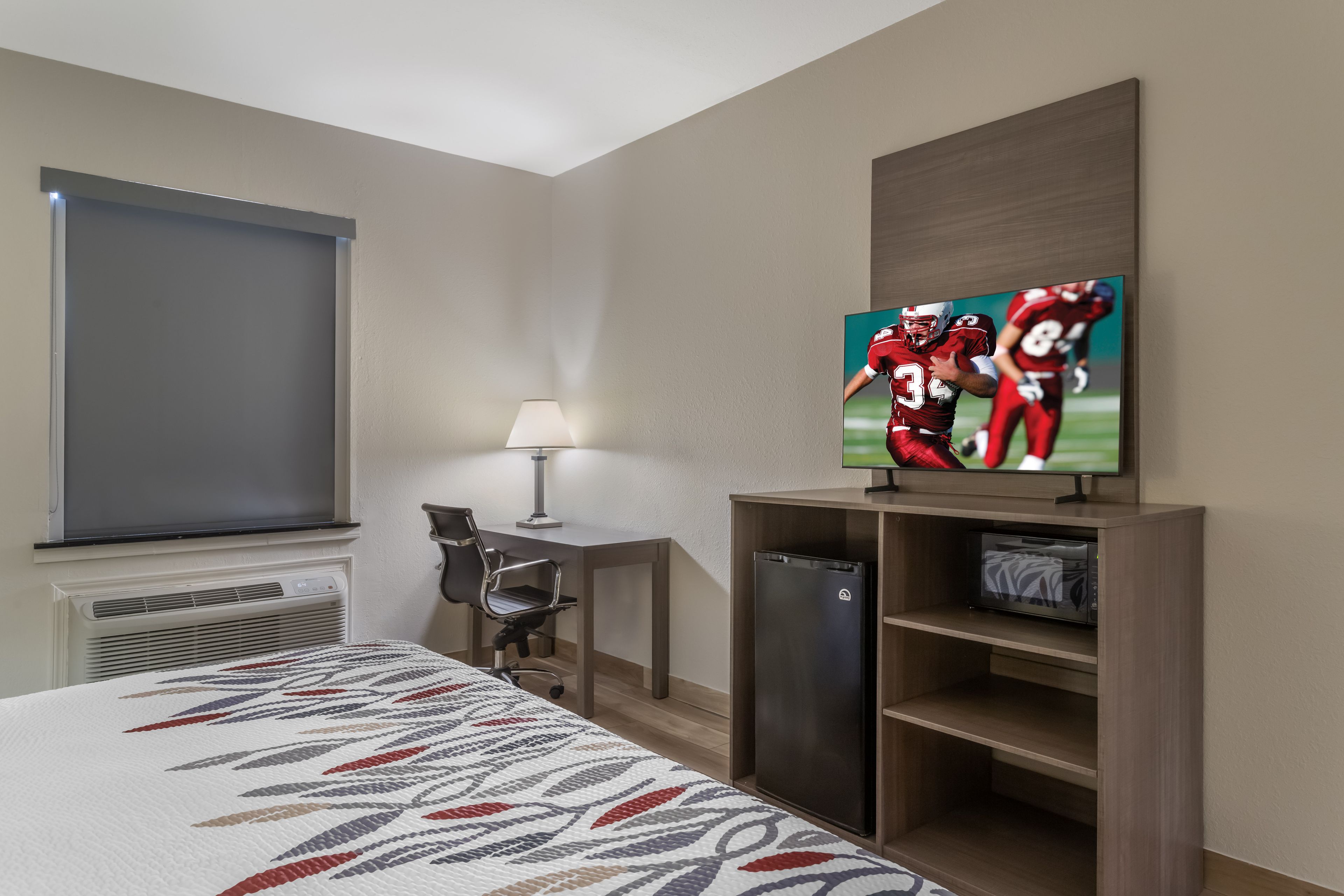Hotelkamer met een bed, bureau, stoel, lamp, televisie en een plank met sportkunst.