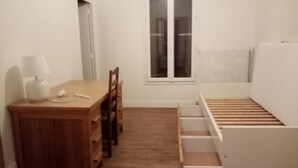 Ein kleiner, schlicht eingerichteter Raum mit einem Holztisch, einem Stuhl, einer Lampe und einem Bett mit Holzgestell.