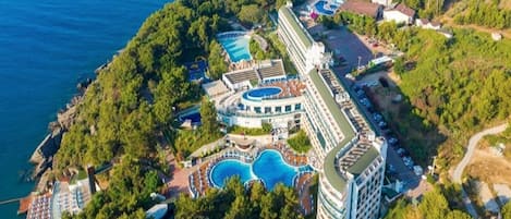 An aerial view of a coastal resort with multiple swimming pools, a tennis court, and a large building with balconies.